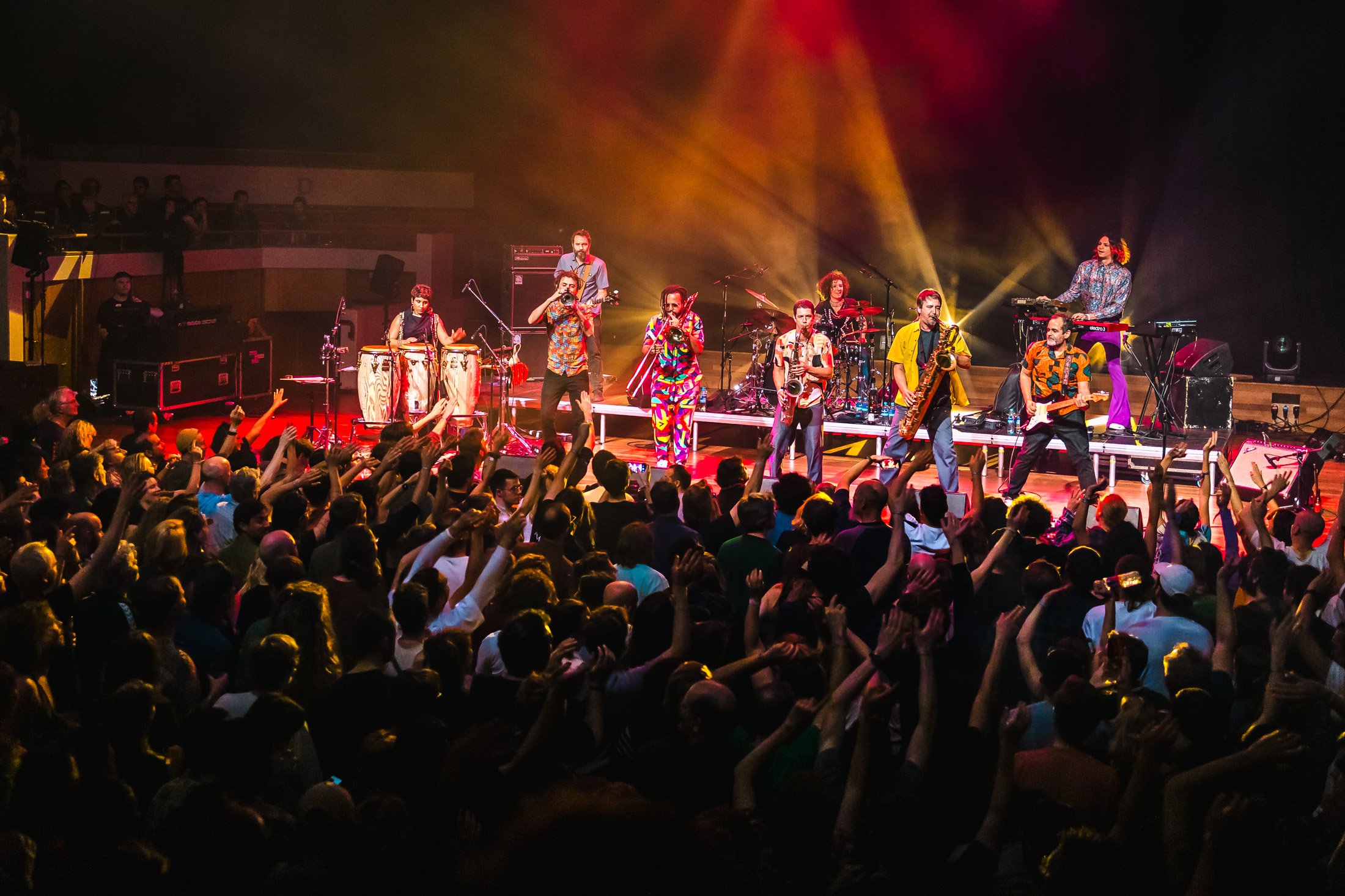 Brazilian band Bixiga 70 during their performance at Le Guess Who Festival, in the Grote Zaal of TivoliVredenburg. The band is on stage performing, while the audience in front of them is dancing and waving